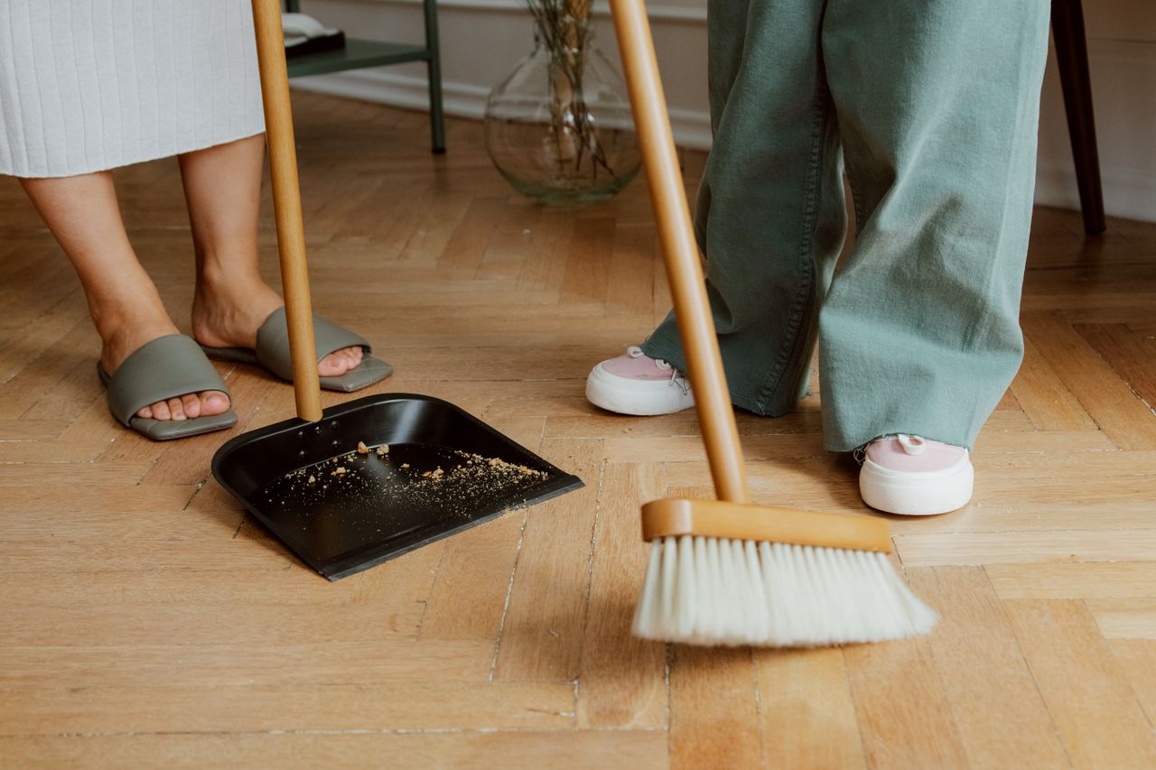 team-02 Close-up of people cleaning wooden floor with broom and dustpan indoors.